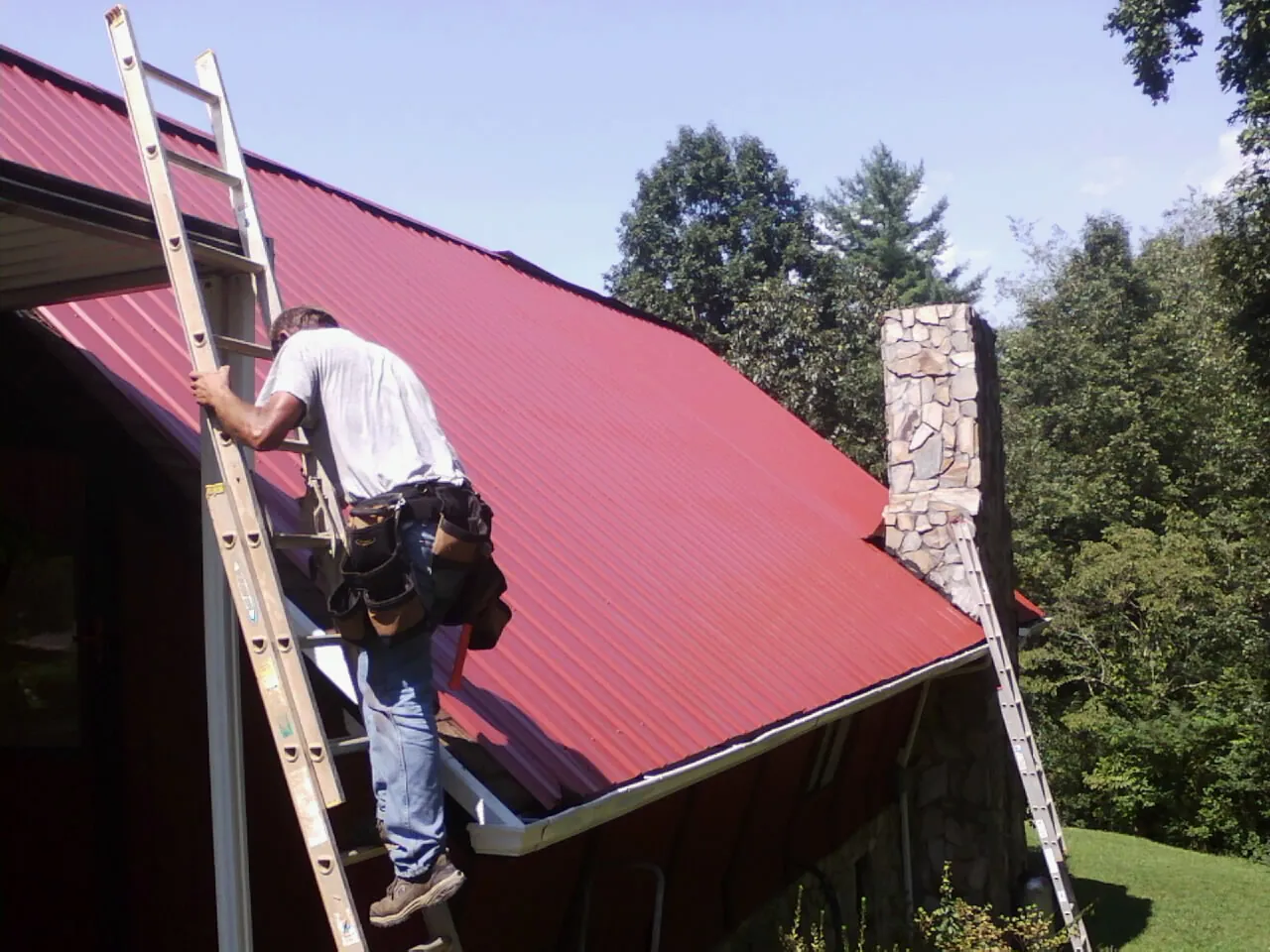 Roofer on a ladder beside a red metal roof and stone chimney — Soffit & Fascia Repair by Roofing Brothers in California